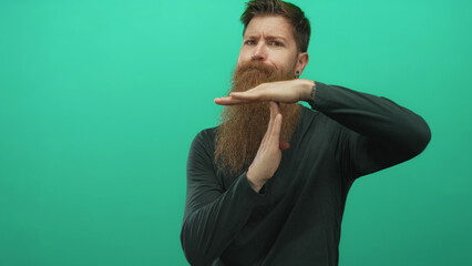Young man with long beard standing makes timeout gesture with hands in green studio setting;...