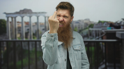 Bearded man in denim jacket flips middle finger with open mouth at ancient roman ruins building;...