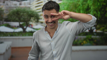 Young hispanic man smiling in an outdoor park, making a peace sign near his eye, surrounded by greenery and bright natural light, capturing a moment of joy and positivity.