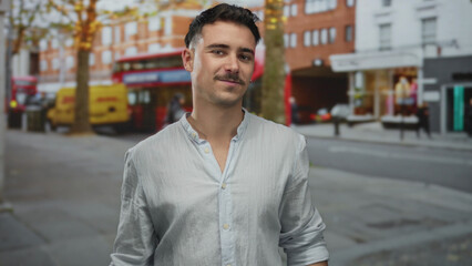 Fototapeta premium Young man with short hair standing on a busy city street with red buses and shops in the background, showcasing urban vibe and relaxed atmosphere.