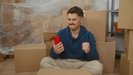 Young man sitting in new home surrounded by boxes, using smartphone with excited expression, in...