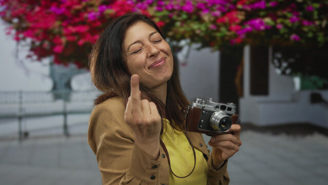 Woman holds vintage camera making middlefinger gesture on street under blooming bougainvillea; defiance rebellion protest resistance.