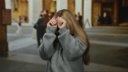 Fototapeta premium Blonde young woman in grey sweater appears scared at an outdoor university setting with old architecture in background showing public area.