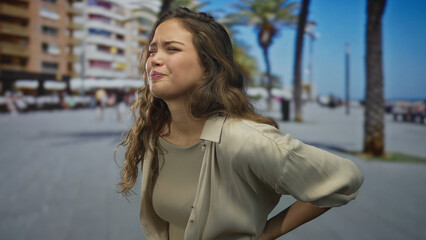 Fototapeta premium Woman expressing pain at seaside promenade outdoors with palm trees, wearing beige blouse, young hispanic highlighting discomfort under bright sunlight.
