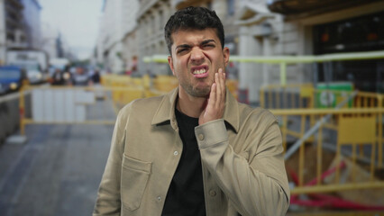 Obraz premium Man presses jaw while touching street fence and young hispanic outdoor male feels ache and discomfort.
