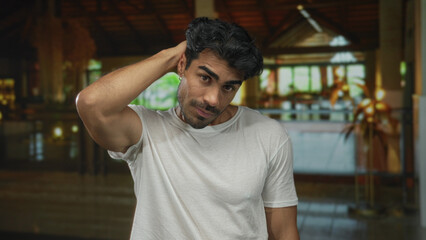 Man in white shirt standing with hand on neck and head slightly tilted down in hotel building;...