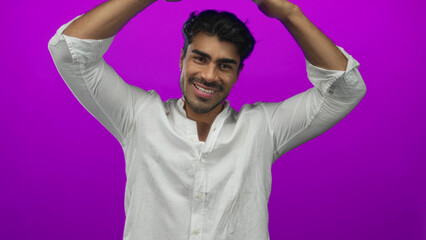 Young hispanic man holds hands in roof shape gesture above his head in studio with purple...