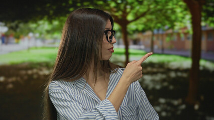 Woman points finger upward in public park near green foliage, wearing glasses and striped blouse;...
