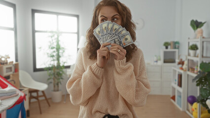 Woman smiling holding us dollar bills in cozy living room with plants and modern furniture,...