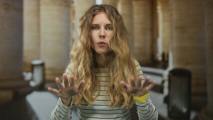Woman raises hands calming in corridor featuring blonde young subject showing deescalation of...