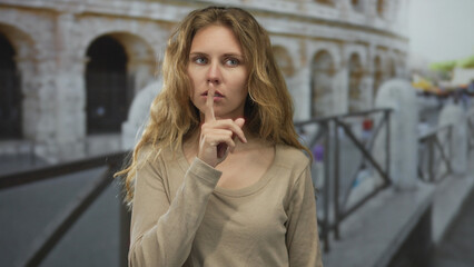 Fototapeta premium Woman raises a finger for a shush moment in italy rome colosseum, as a blonde young traveler presses lips and flips hair in a casual portrait.