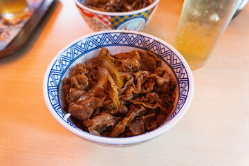 A close-up of a delicious bowl of Japanese gyudon. Thinly sliced beef and onions simmered in savory sauce over rice, served with chopsticks and iced tea on a wooden table.