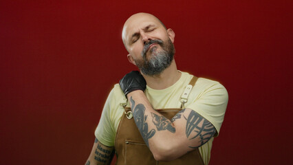 Man with beard wearing apron over red background touching his neck appearing in pain isolated in studio setting.