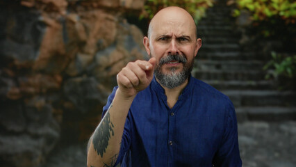 Bald man with a beard in blue shirt gestures seriously in an outdoor urban setting with a rocky...