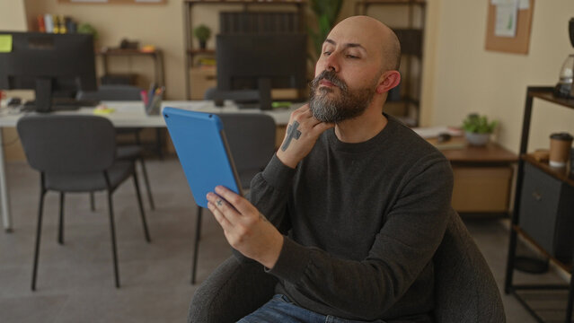 Bald man using tablet in cozy office with modern decor and workspace essentials around, showcasing professional and focused atmosphere.
