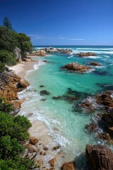 Fototapeta premium Tranquil coastal scene. Turquoise water laps sandy beach, surrounded by rocky outcrops. Lush green vegetation borders the shoreline. Bright, sunny day