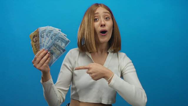 Young woman with blonde hair excitedly holding hungarian forint banknotes against a blue background, expressing surprise and delight with wide eyes and open mouth.