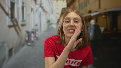 Young woman in red volunteer shirt brings hand to mouth to whisper on cobblestone street; secrecy...