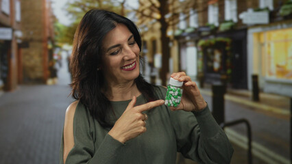 Hispanic woman in green sleeveless top points finger to clear pill bottle of green and white capsules while smiling on a sunlit street with storefronts; wellness.