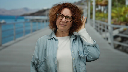 Woman gesturing on seaside promenade with ocean and mountains in the background, wearing glasses...