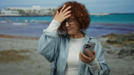 Woman on beach holding smartphone appears frustrated while enjoying seaside view, dressed casually...
