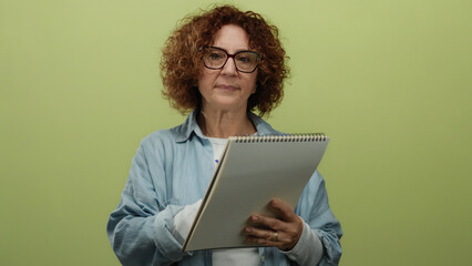 Middle-aged woman writing in notebook against isolated yellow background, wearing glasses and...