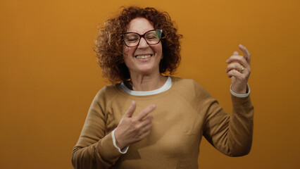 Middle-aged hispanic woman playfully mimics playing guitar against an isolated orange wall...