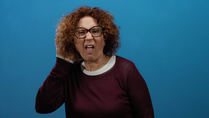 Middle-aged hispanic woman with curly hair and glasses gestures as if listening on an isolated blue...