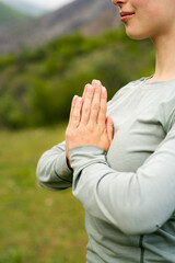 Meditation in nature. Smiling friendly woman meditating in green mountains, holding her hands in namaste gesture, wearing long sleeve, close-up outdoor yoga activities in summer