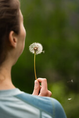 Vertical close-up of young woman blowing dandelion seeds with green blurred background, girl holding dandelion seed ball and making wish, happiness, desire, hope, dream concept
