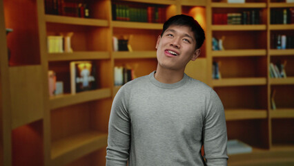 Young man in library with shelves in the background displaying a playful expression in an indoor room setting, showcasing a contemporary casual style.