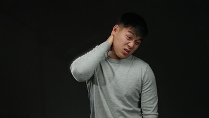 Young chinese man in grey sweater stands against a black background expressing discomfort touching...