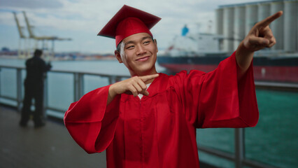 Young man in red graduation gown and cap celebrates on a boat cruise pointing towards the seaside...