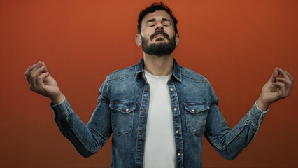 Hispanic man meditating with a beard and denim jacket against an orange background, hands in prayer...