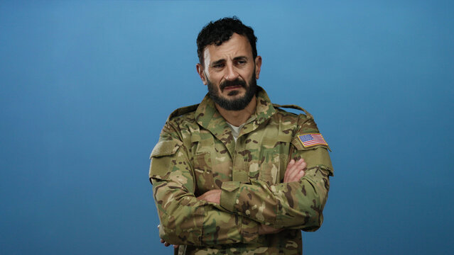 Hispanic man with beard in military uniform stands against blue background displaying serious expression and subtle defiance.