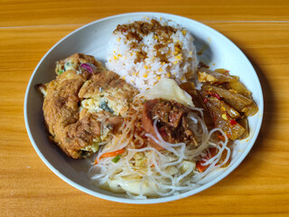 A typical Indonesian mixed rice dish with white rice sprinkled with spices, omelet, vermicelli, boiled vegetables and other side dishes on a white plate. Wooden table background.