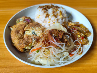 A typical Indonesian mixed rice dish with white rice sprinkled with spices, omelet, vermicelli, boiled vegetables and other side dishes on a white plate. Wooden table background.