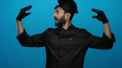 Hispanic man with beard in black chef uniform and gloves making expressive gestures against blue...