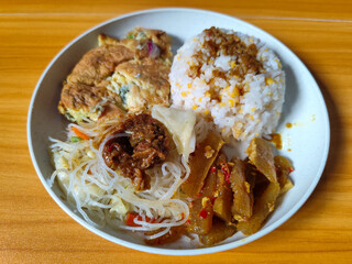 A typical Indonesian mixed rice dish with white rice sprinkled with spices, omelet, vermicelli, boiled vegetables and other side dishes on a white plate. Wooden table background.