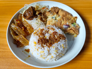 A typical Indonesian mixed rice dish with white rice sprinkled with spices, omelet, vermicelli, boiled vegetables and other side dishes on a white plate. Wooden table background.