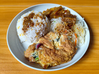 A typical Indonesian mixed rice dish with white rice sprinkled with spices, omelet, vermicelli, boiled vegetables and other side dishes on a white plate. Wooden table background.