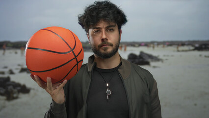 Young man with beard holds basketball at a seaside beach, showcasing enjoyment of outdoor sports...
