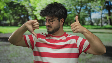 Young man with beard in striped shirt making thumbs up and down gestures in a green park outdoors...