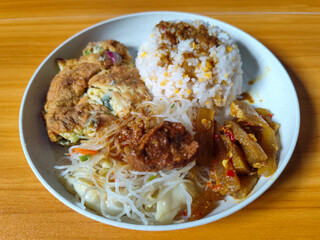 A typical Indonesian mixed rice dish with white rice sprinkled with spices, omelet, vermicelli, boiled vegetables and other side dishes on a white plate. Wooden table background.