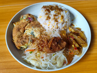 A typical Indonesian mixed rice dish with white rice sprinkled with spices, omelet, vermicelli, boiled vegetables and other side dishes on a white plate. Wooden table background.