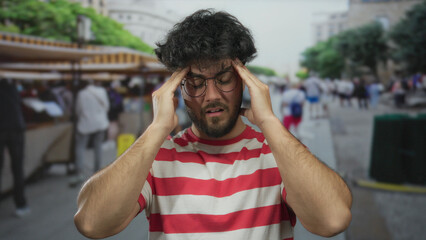 Young hispanic man with a beard wearing glasses and red-striped shirt appears stressed outdoors on...