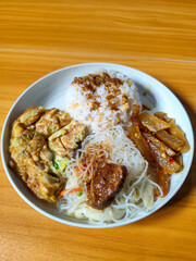 A typical Indonesian mixed rice dish with white rice sprinkled with spices, omelet, vermicelli, boiled vegetables and other side dishes on a white plate. Wooden table background.