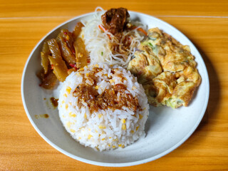 A typical Indonesian mixed rice dish with white rice sprinkled with spices, omelet, vermicelli, boiled vegetables and other side dishes on a white plate. Wooden table background.