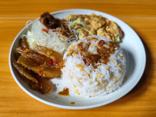 A typical Indonesian mixed rice dish with white rice sprinkled with spices, omelet, vermicelli, boiled vegetables and other side dishes on a white plate. Wooden table background.