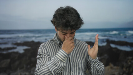 Young man with beard holding nose outdoors at the seaside due to bad smell while standing by the...